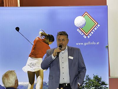 A man in a suit speaks into a microphone in front of a golf banner showing a woman swinging a club, promoting golf in Austria, with mountains in the background.