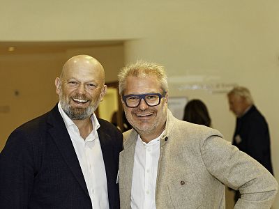 Zwei lächelnde Männer in Anzügen posieren bei einer Veranstaltung. Der Raum ist gut beleuchtet, im Hintergrund sind unscharfe Personen zu erkennen.