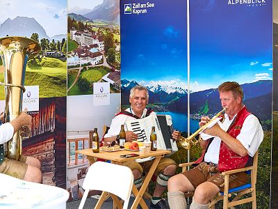 Three musicians in traditional Austrian attire play brass and accordion instruments in front of scenic mountain and countryside posters.