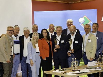 Gruppe von Geschäftsleuten bei einer Veranstaltung posieren vor einem Bildschirm. Zwei Männer halten Trophäen. Einige tragen Namensschilder.