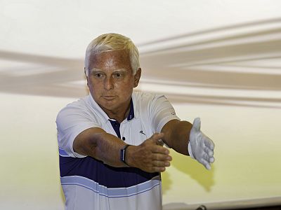 An older man with white hair demonstrates a golf swing. He wears a white and blue polo and a golf glove on one hand against a light background.