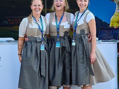 Three women in traditional Bavarian dirndls, smiling and posing at an event, wearing lanyards and name tags, against a background of outdoor scenery.