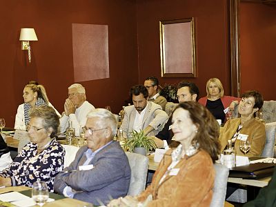 Several people sit in a conference room with red walls, listening attentively. Some are taking notes. The atmosphere is professional and focused.