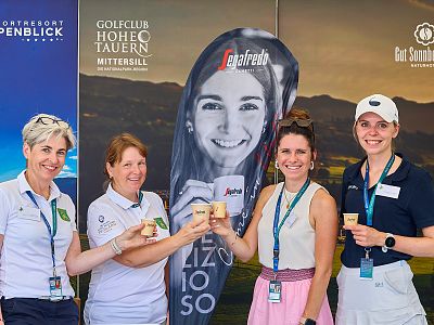 Four women smiling and holding drinks at a golf event in front of banners displaying golf club logos.