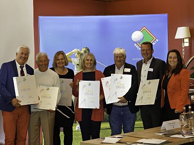 Seven people holding certificates stand in a row, smiling at an indoor event. They appear to be celebrating an achievement or recognition.