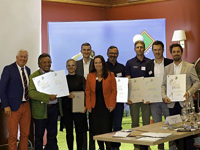 A group of smiling people standing together in a room, holding certificates and awards, with a blue and green backdrop behind them.
