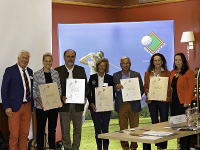 A group of people standing indoors holding certificates, smiling at the camera. They are dressed in formal attire with a golf-themed backdrop behind them.