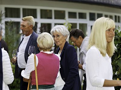 People in formal attire standing and conversing at an outdoor event with wine glasses, against a backdrop of a modern building and greenery.
