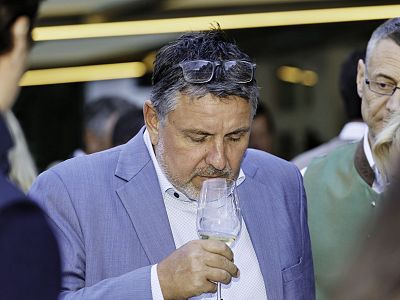 Man in suit smells a glass of white wine at an event, surrounded by other people, creating a formal atmosphere.