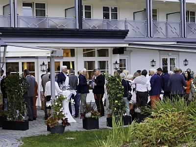 People gathered outdoors at an elegant venue, dressed formally, engaging in conversation during a social event. The setting includes plants and a sophisticated building.