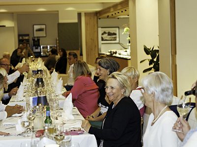 A group of people seated at a long dining table in a restaurant setting. The table is elegantly set with glasses and napkins. The atmosphere is lively.