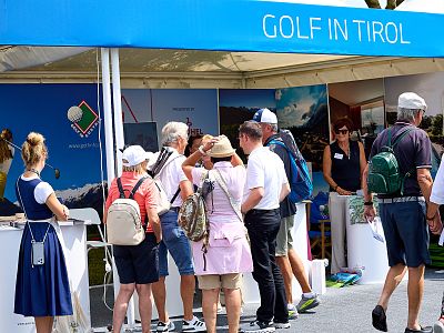 A group of people stand near a booth labeled 'Golf in Tirol' with promotional materials. Some wear hats and bags. The booth has a blue canopy.