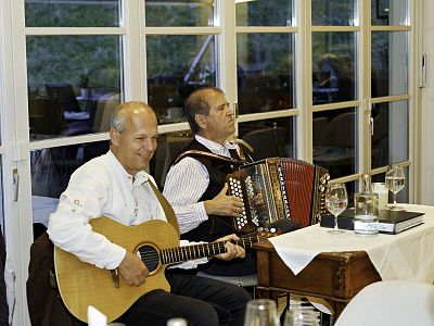 Two musicians, one with a guitar and the other with an accordion, perform at a table in a restaurant with white tablecloths and glassware.