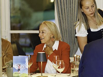 Two women seated at a table during a dinner. One in a red blazer smiles, while the other in a traditional outfit stands nearby. The table is elegantly set.
