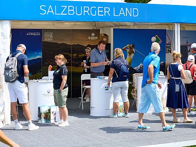 People gather at a Salzburger Land promotion booth outdoors. They look at brochures and engage with staff under a blue and white tent.