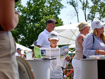 A young boy reads a booklet at an outdoor event, surrounded by adults in casual attire. Umbrellas and trees indicate a sunny day with people chatting and walking.