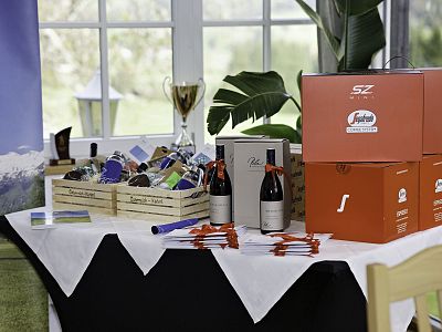 Table with wine bottles, gift boxes, and small trophies arranged against a window with lush greenery outside, suggesting a celebratory occasion.