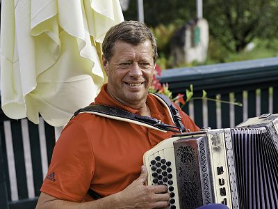 Man in red shirt playing an accordion outside with a white umbrella and greenery in the background.