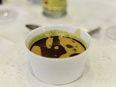 White bowl with soup garnished with dark sauce swirls on a white tablecloth; blurred utensils in background suggest a dining setup.