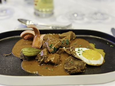 A plated dish with meat slices in brown sauce, a sunny-side-up egg, pickles, and a vegetable garnish on an elegant platter.