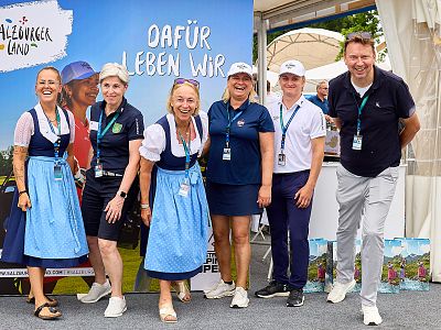 Six people posed together at an event with a backdrop reading 'DAFÜR LEBEN WIR.' Two wear traditional blue dresses, others wear casual attire. Outdoor setting.