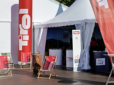 Outdoor exhibition booth with Tirol branding, deck chairs, and promotional banners in a sunny setting.