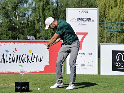 Golfer in action on the course, mid-swing, during the Austrian Alpine Open with sponsor signs and greenery in the background.