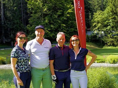 Vier Personen stehen nebeneinander auf einem Golfplatz mit Bäumen im Hintergrund. Alle tragen sportliche Kleidung und lächeln in die Kamera.