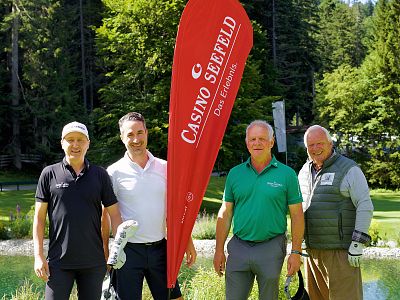 Four men stand on a golf course near a red banner that reads 'CASINO SEEFELD'. Behind them are lush green trees under a sunny sky.