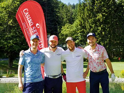 Four men on a golf course, smiling with a Casino Seefeld flag in the background, surrounded by trees.