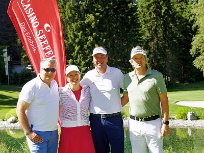 Four people are smiling on a golf course, wearing casual golf attire. A red banner and trees are visible in the background.