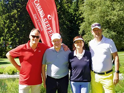 Four people stand on a golf course, smiling at the camera. They're casually dressed, with a red banner and trees in the background.