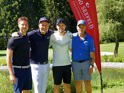Vier Männer stehen lächelnd nebeneinander auf einem Golfplatz vor einem Teich, umgeben von Bäumen. Sie tragen Golfkleidung und eine rote Fahne weht im Hintergrund.