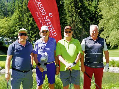 Vier Männer stehen auf einem Golfplatz vor einem roten Banner, jeder hält einen Golfschläger. Der Hintergrund zeigt grüne Bäume und einen klaren, blauen Himmel.