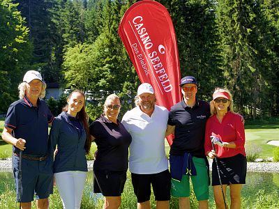 A group of six people smiling, standing with a red flag on a golf course, surrounded by greenery and trees, on a sunny day.