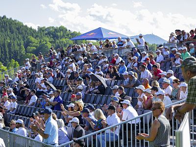 Crowd seated in bleachers outdoors under blue sky with trees in background, wearing hats and sunglasses, attending an event on a sunny day.