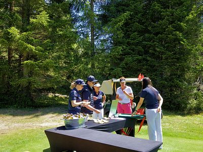People gather around a table in a forest area, enjoying a sunny day. Trees tower in the background, and casual summer attire is worn by all.