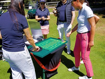 Four people outside on grass, standing around a small casino game table, engaged in play. Sunny day with casual clothing and green surroundings.