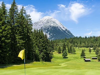 A scenic golf course surrounded by evergreen trees with a snow-capped mountain in the background and a yellow flag marking the green.