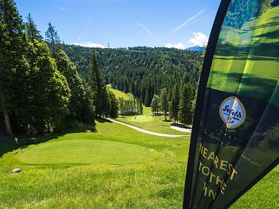 Golf course surrounded by dense forest, with green fairway and flag in foreground, under a blue sky with scattered clouds.