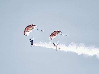 Two parachutists with red and white parachutes perform stunts in a clear sky, leaving white smoke trails behind them.