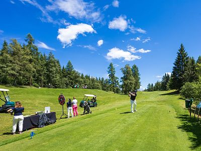 Golfers in action on a green course with trees and blue sky, using golf carts and equipment.