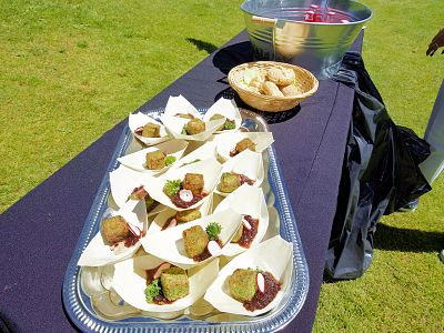 Appetizers in paper cups on a tray with rolls in a basket nearby on a black tablecloth outdoors.