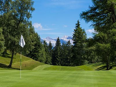 A golf course with a flagpole on a green, surrounded by trees and distant snow-capped mountains under a clear sky.
