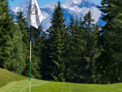 Golf flag on a green course surrounded by pine trees and mountains in the background. The sky is clear, highlighting the scenic beauty of the landscape.