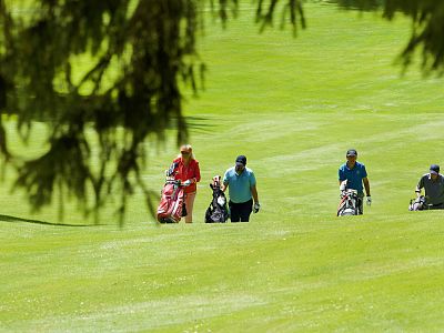 Four golfers walk across a green golf course, pushing trolleys. The foreground shows tree branches, and the sun illuminates the lush grass.