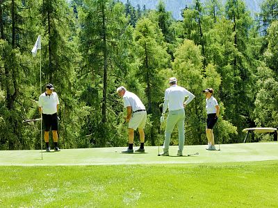 Four people stand on a golf putting green surrounded by lush trees. One is putting as others watch, enjoying a game on a sunny day in a wooded area.