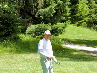 Ein älterer Mann steht auf einer grünen Wiese, umgeben von hohen Bäumen. Er trägt eine weiße Mütze und ein weißes Outfit. Es scheint ein sonniger Tag zu sein.