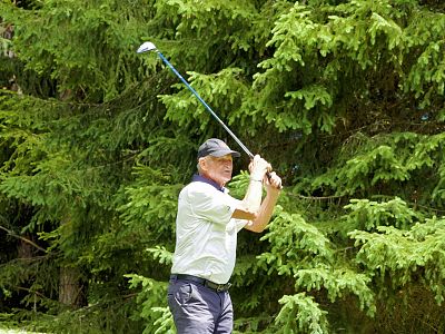 A man in a white shirt and cap is swinging a golf club on a course, surrounded by green trees.