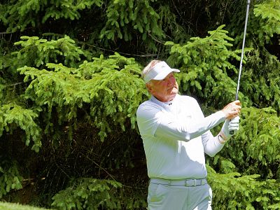 A person in a white outfit is swinging a golf club surrounded by green trees, indicating they are playing golf outdoors.